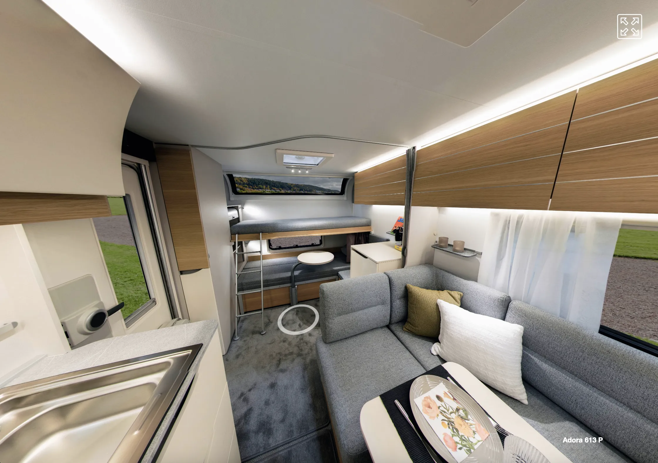Asuntoauton sisätila, jossa on harmaa sohva, puinen säilytystila ja kerrossänky, valaistus on kirkas ja moderni.