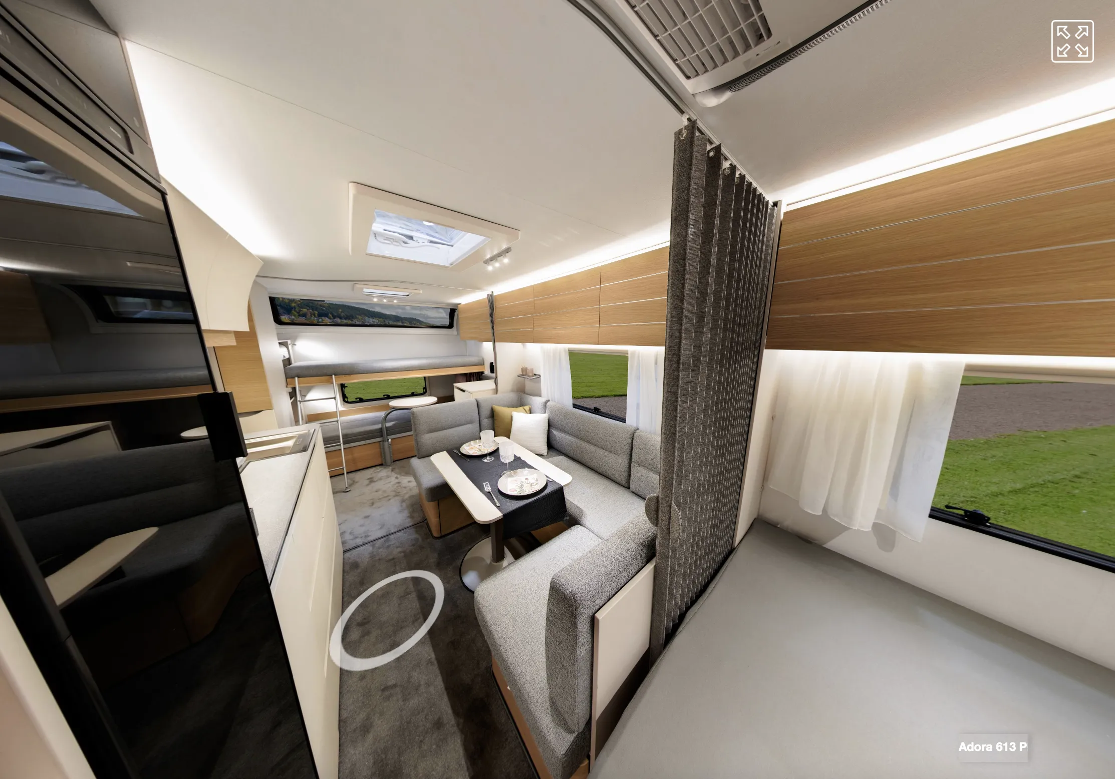 Asuntoauton sisätila, jossa on harmaat sohvat, puinen ruokapöytä ja valkoiset verhot, valaistus on kirkas.