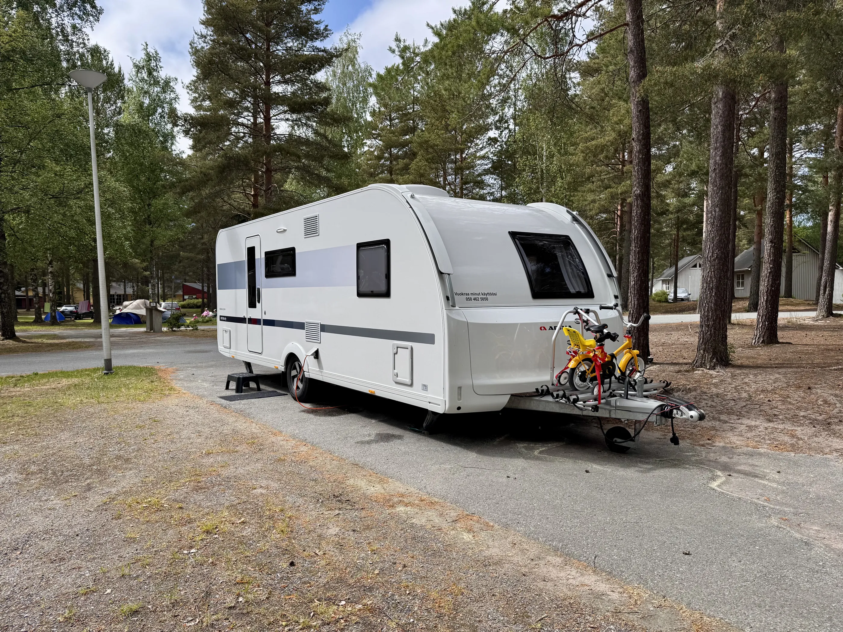 Valkoinen asuntovaunu pysäköitynä leirintäalueella, jossa on polkupyöräteline ja kaksi lasten polkupyörää. Taustalla näkyy puita ja muita leirintäalueen rakennuksia. Valoisa ja aurinkoinen sää.