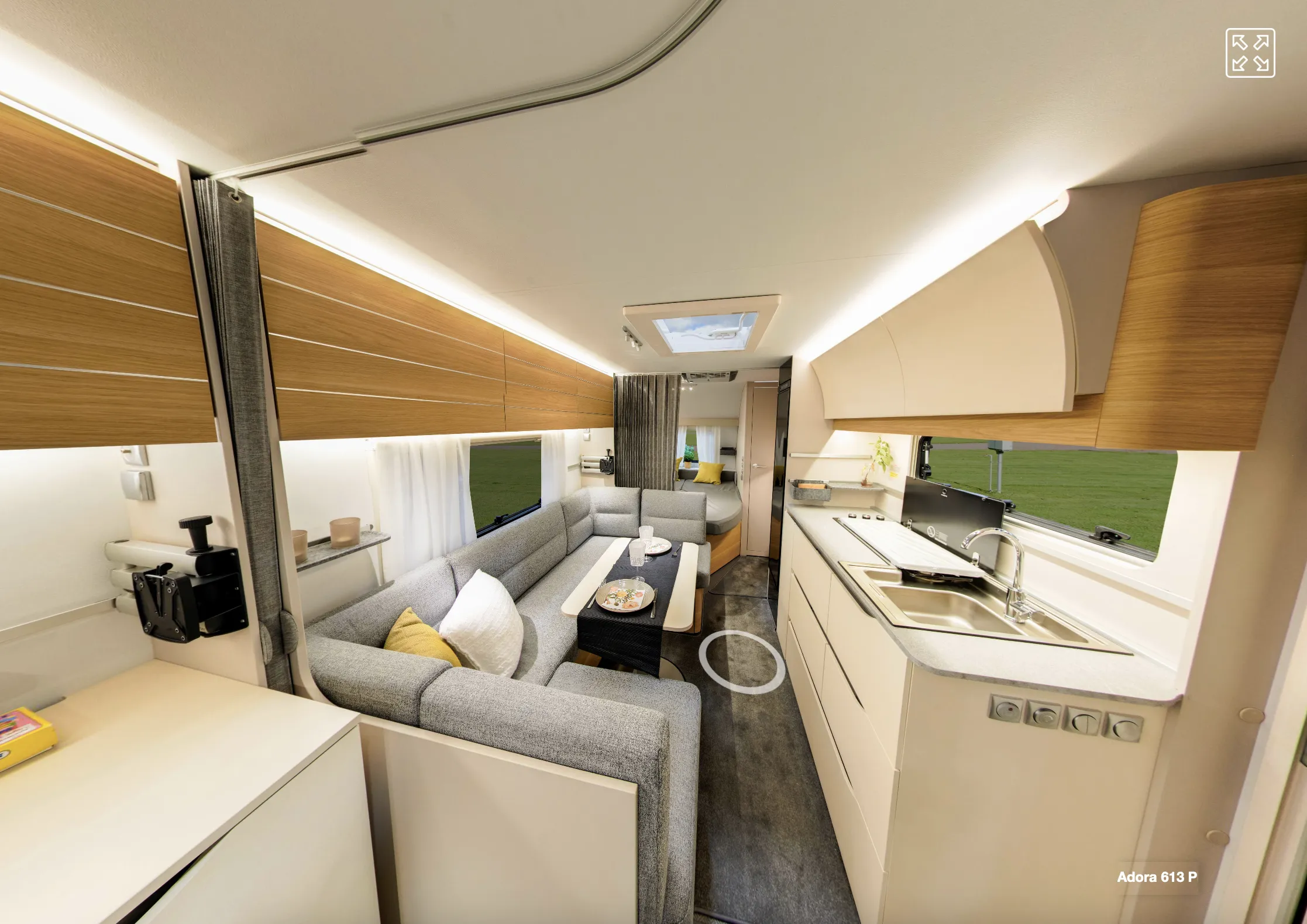 Panoraamanäkymä asuntovaunun sisätiloista, jossa on harmaa sohva, puinen säilytystila ja valoisa keittiö.