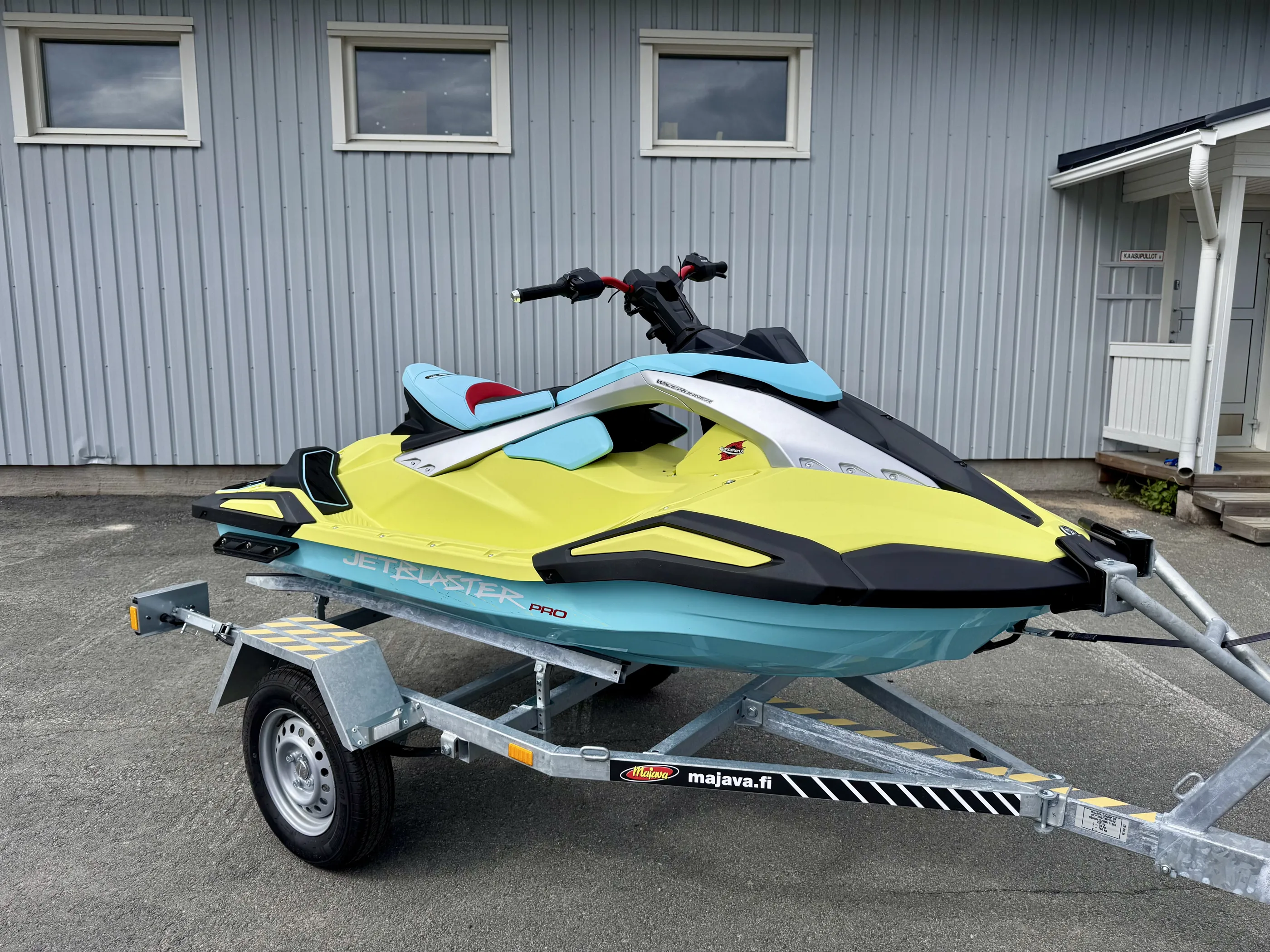 Keltainen ja turkoosi Yamaha JetBlaster Pro -vesijetti harmaalla peräkärryllä harmaan rakennuksen edessä, jossa on kolme ikkunaa.