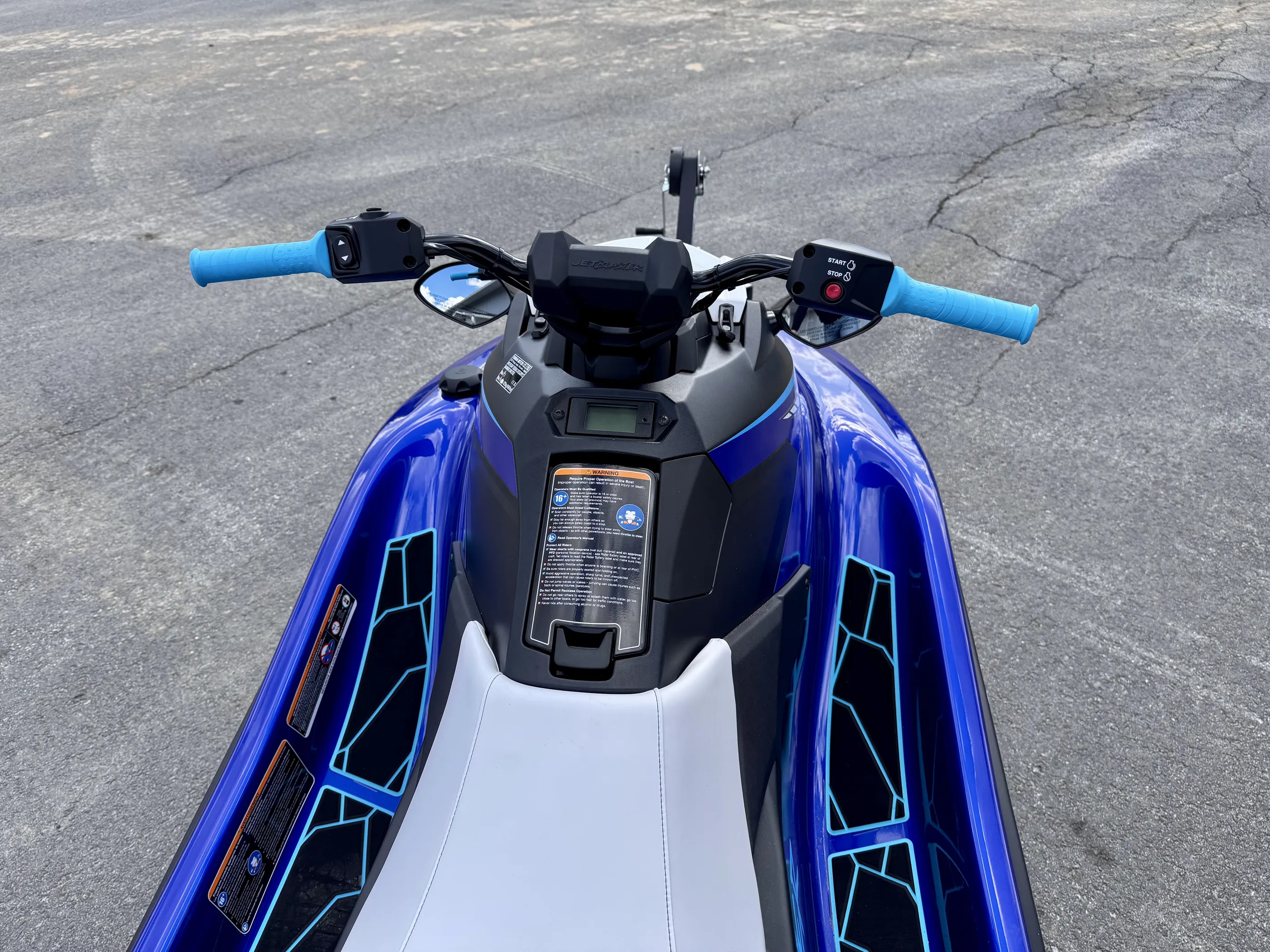 Lähikuva sinisestä vesiskootterista, jossa on vaaleansiniset kädensijat, valkoinen istuin ja musta ohjauspaneeli. Varoitustarra näkyy selvästi.