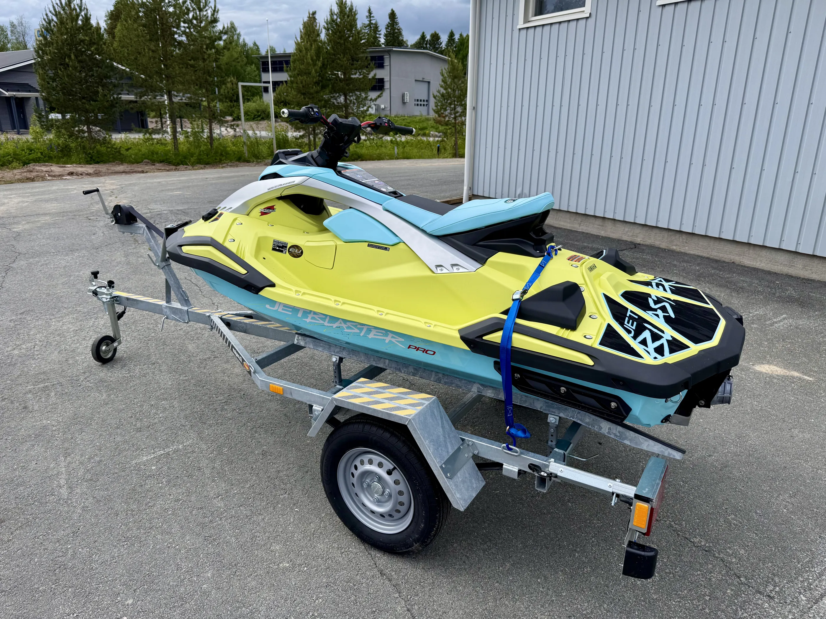 Keltainen ja sininen vesiskootteri peräkärryllä asfalttipinnalla, taustalla rakennuksia ja puita. Valoisa sää.