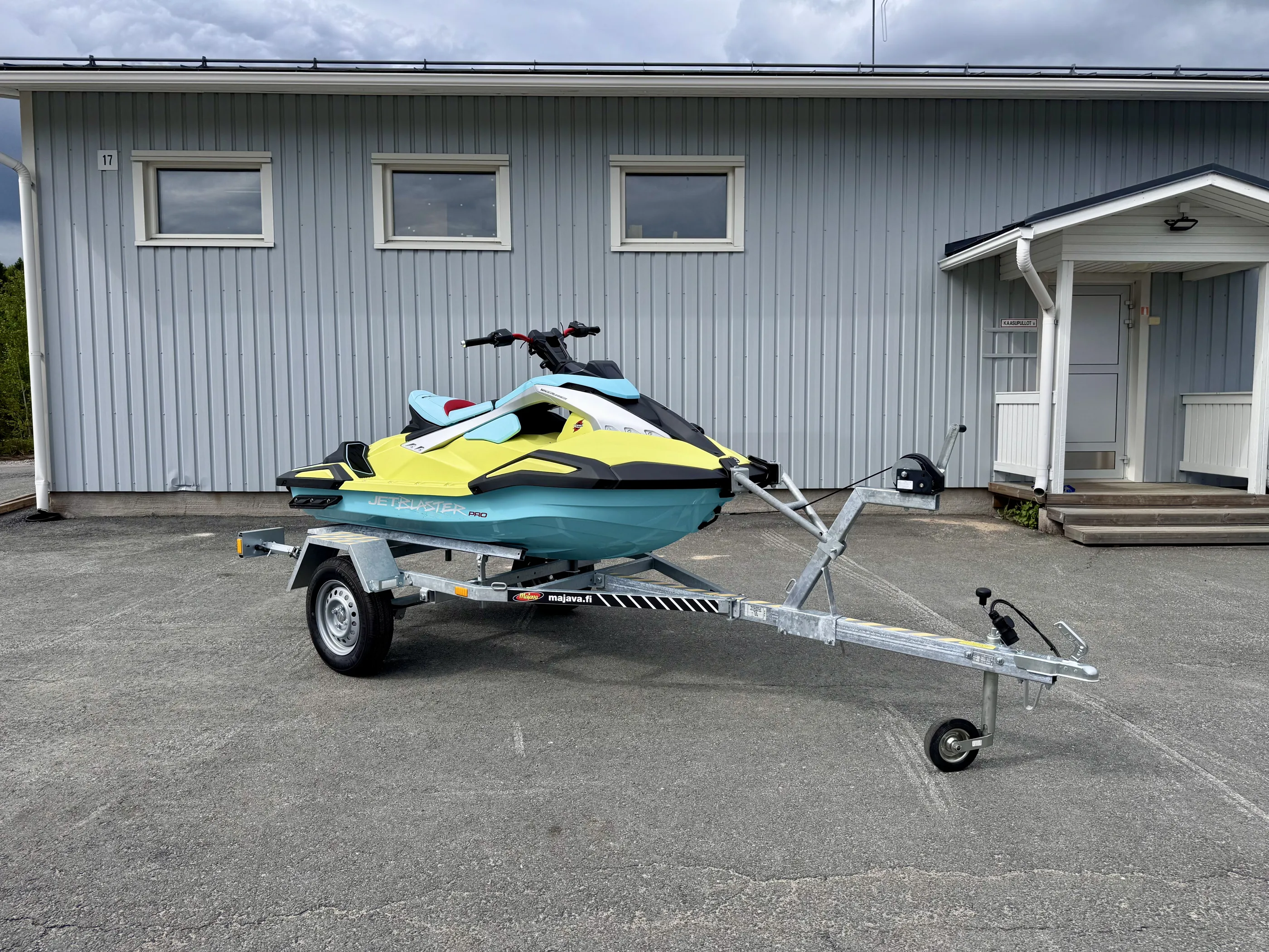 Vaaleansininen ja keltainen vesiskootteri trailerilla harmaalla pihalla harmaan rakennuksen edessä, pilvisellä säällä.