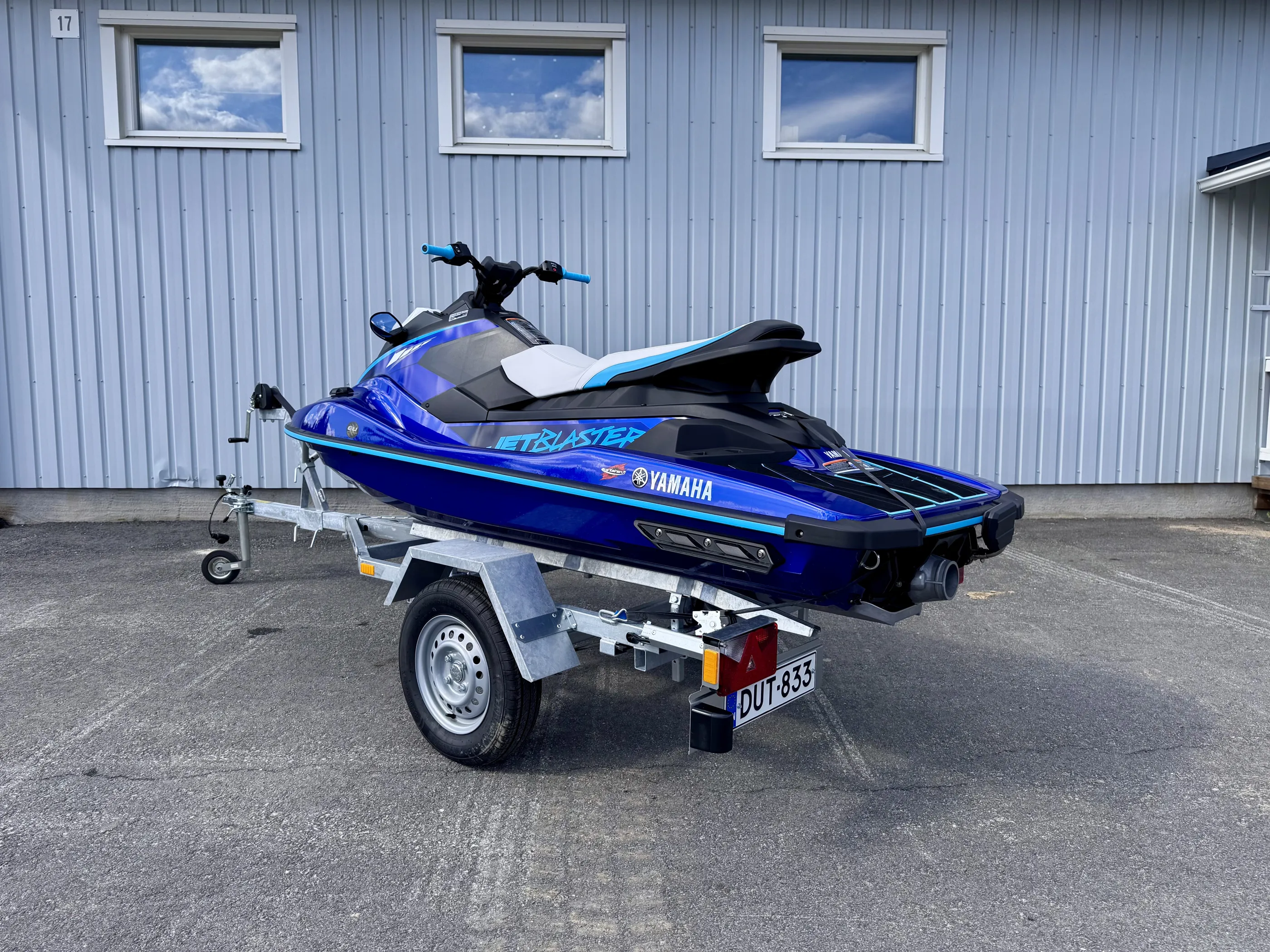 Sininen Yamaha Jet Blaster -vesiskootteri trailerilla harmaata seinää vasten, jossa on kolme ikkunaa ja numero 17. Valoisa päivä.
